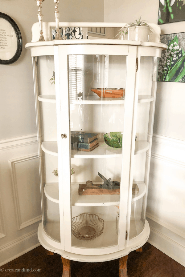 A white curio cabinet with glass in the corner of a room with décor pieces on the shelves.