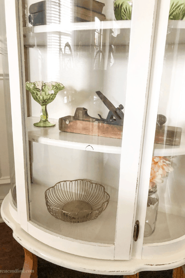 A painted white curio cabinet with glass and décor pieces on the shelves.