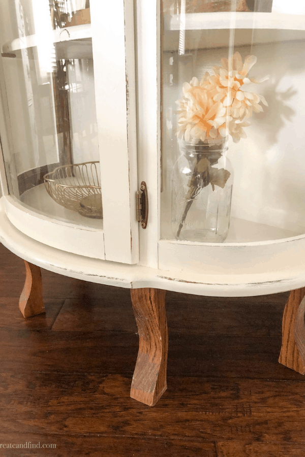 The bottom of a white painted curio cabinet with wooden legs and décor pieces behind the glass.