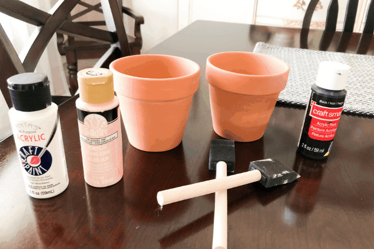 Orange clay pots with a bottle of paint and two foam paintbrushes on a dining room table.