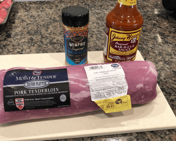 A bottle of BBQ sauce, a bottle of BBQ seasoning, and a package of pork tenderloin on a cutting board on a granite countertop.