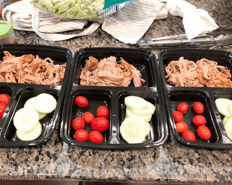 Shredded pork with tomatoes and sliced cucumbers in black meal prep containers on a kitchen counter.