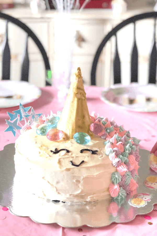 A birthday cake with a gold unicorn horn and smiley face on a table with a pink tablecloth and party plates.