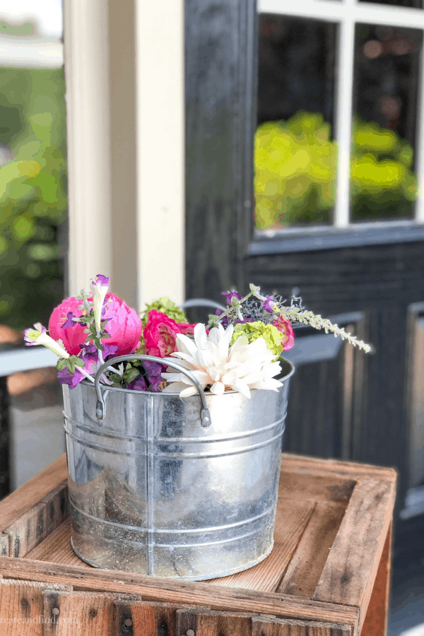 White pink and purple faux flower arrangement in a metal container on a porch.