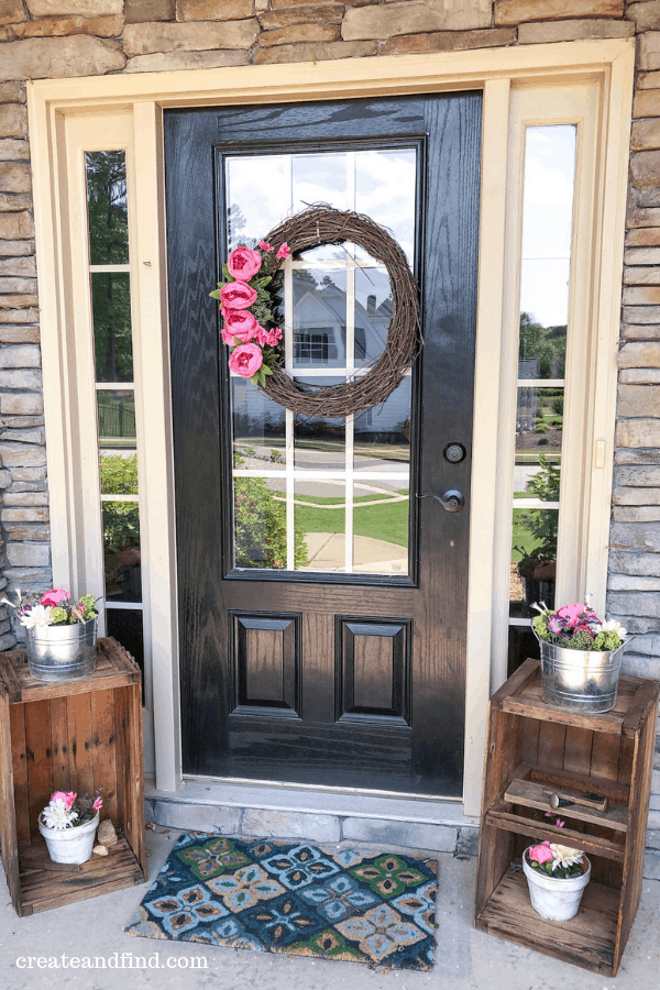 Summer front porch décor including a wreath with pink flowers hanging from the door and metal tins with faux flower arrangements.