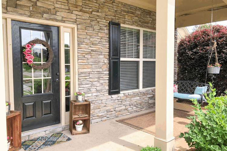 Front porch with stone walls, a black door with a wreath, and rugs.