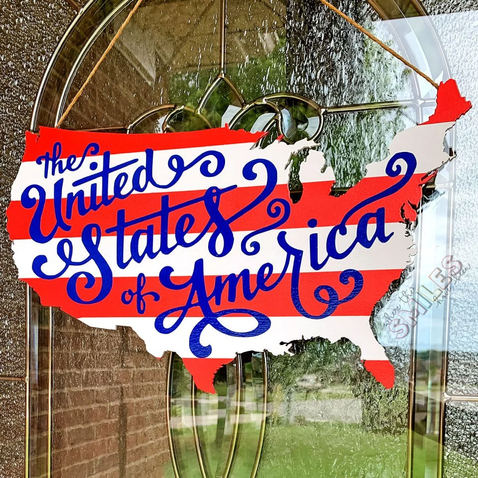 A wooden front door hanger in the shape on the Unite states with red and white stripes and blue text that reads "The United States for America" in script font.
