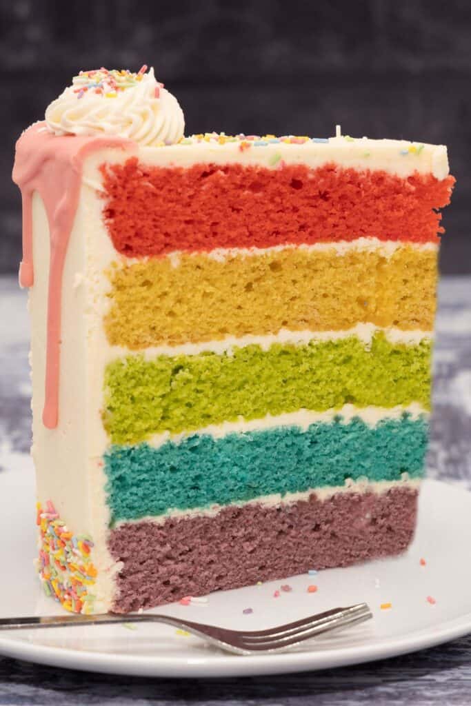 A large slice of cake with the inside layers exposed in the colors of the rainbow.