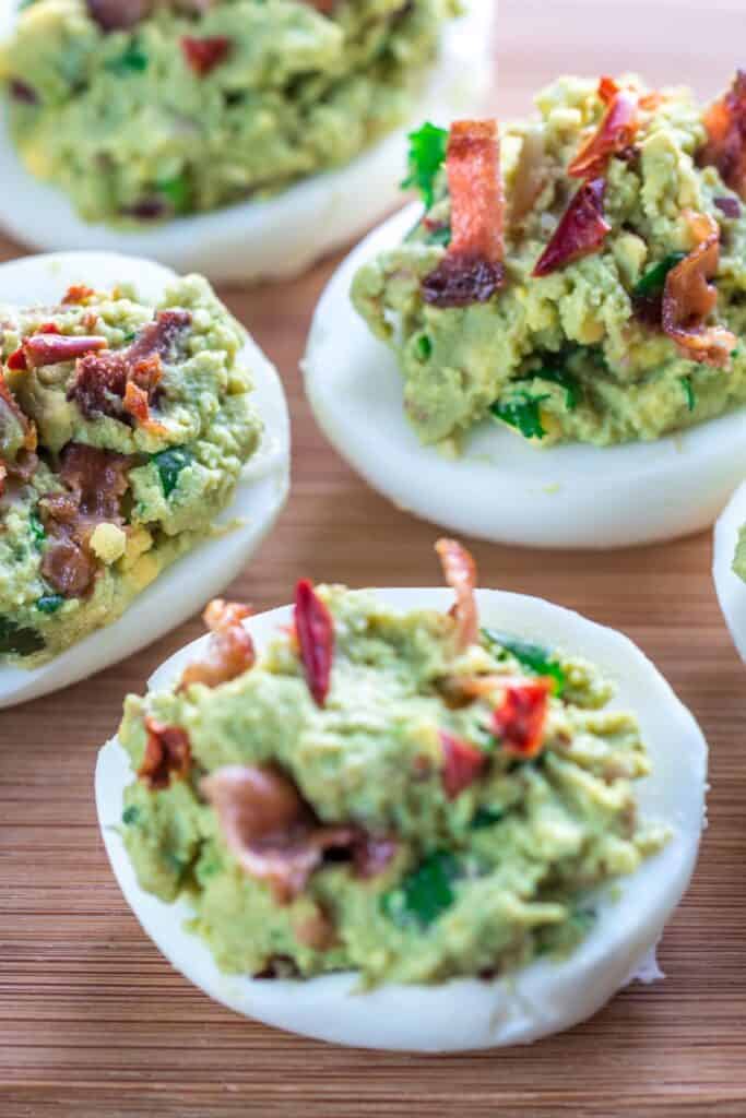 Green deviled eggs with avocado and bacon on a wooden cutting board.