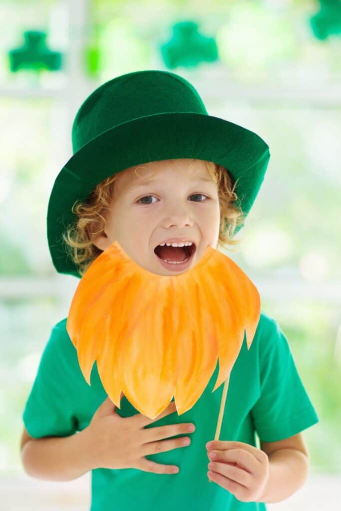 A toddler boy wears a green hat and an orange paper beard.