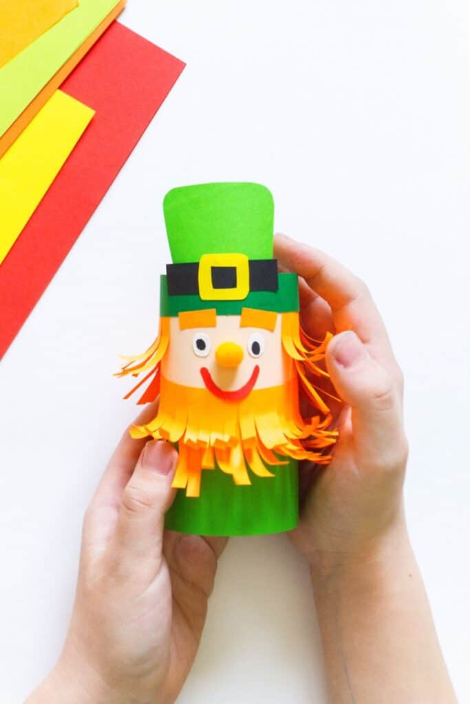 A child holds a leprechaun craft project next to a stack of construction paper.