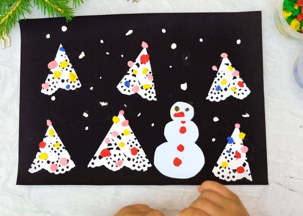 A child makes white trees and and snowman using crafts supplies and paper.-