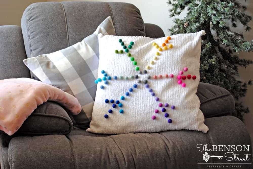 A pom pom rainbow snowflake pillow on a gray couch.