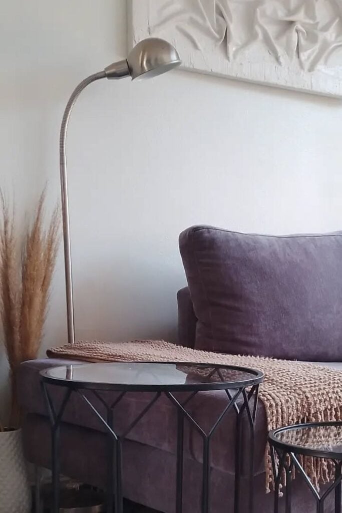 A living room side table next to a couch with a modern lamp, texture wall décor above, and faux grass décor behind.